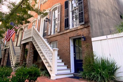A charming brick building with a white staircase, blue door, and American flag, surrounded by greenery.