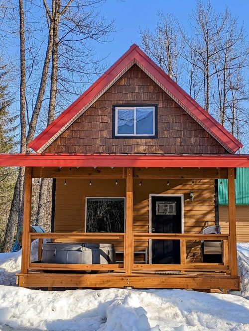 A cozy cabin with a red roof and wooden porch, surrounded by snow and trees under a clear blue sky.