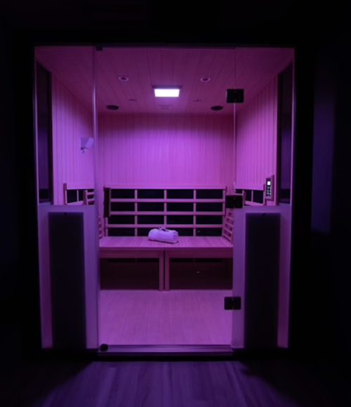 A dimly lit sauna with pink lighting, featuring wooden benches and a glass door.