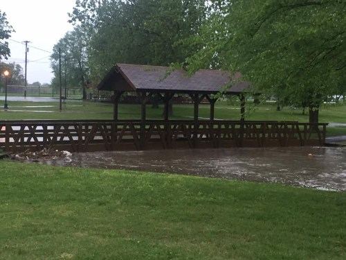 A wooden bridge over a flooded area in a park, with trees and a shelter in the background on a rainy day.