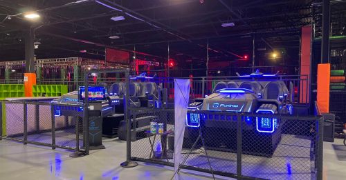 A brightly lit indoor entertainment area featuring futuristic gaming stations with blue lighting and a fenced-off section.