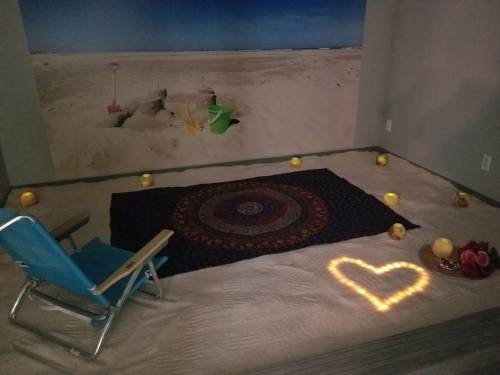 A cozy space with a patterned rug, beach chairs, candles, and a heart shape made of lights on sandy flooring.