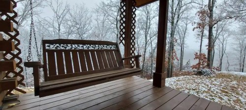 A wooden swing on a porch, surrounded by snowy trees and a misty landscape in the background.