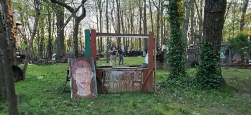A rustic outdoor scene with a wooden booth displaying a sign that says "Sorry, We're Open," surrounded by trees.
