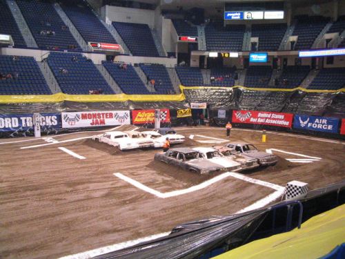 A dirt arena with several wrecked cars arranged for a monster truck event, surrounded by banners and empty seats.