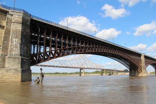 A large metal bridge spans a river, with a smaller bridge visible in the background under a blue sky with clouds.