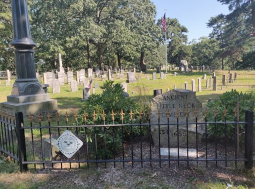 A view of Ancient Little Neck Cemetery, featuring gravestones and a flag in the background, surrounded by greenery.
