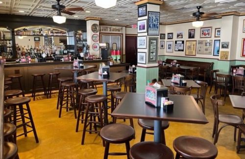 A cozy restaurant interior with wooden tables, bar stools, and a well-stocked bar, featuring framed photos on the walls.