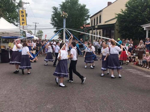 A group of children in traditional costumes dance around a maypole during a street festival, with spectators watching.