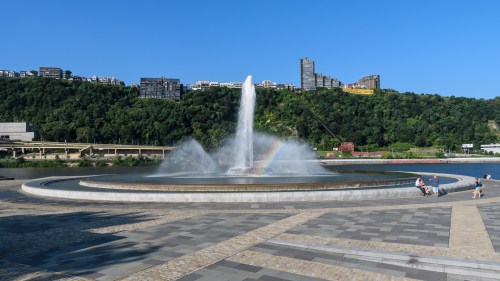 A large fountain sprays water into the air, with a rainbow visible, set against a green hillside and clear blue sky.