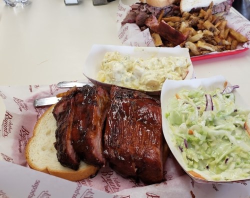 A plate featuring BBQ ribs on bread, sides of coleslaw and macaroni salad, with fries in the background.