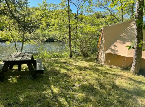 A serene campsite by a river, featuring a tent and a picnic table surrounded by lush greenery and sunlight.