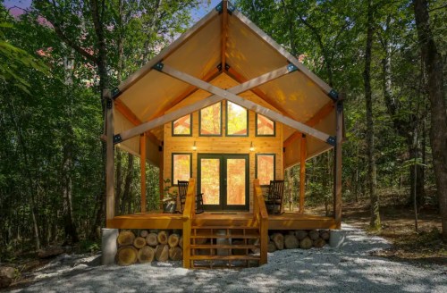 A cozy cabin with a large front porch, surrounded by trees and featuring large windows that reflect autumn colors.