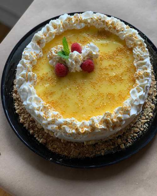 A round cake topped with yellow glaze, whipped cream, raspberries, and golden sprinkles on a crumbly base.