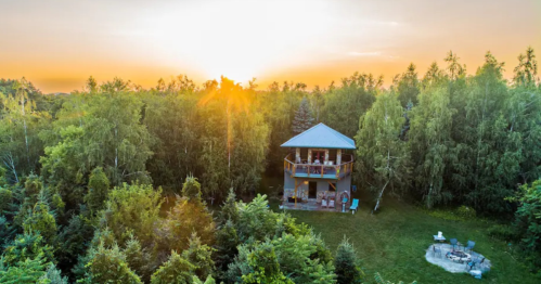 A cozy house surrounded by trees at sunset, with a fire pit in the foreground and warm light illuminating the scene.