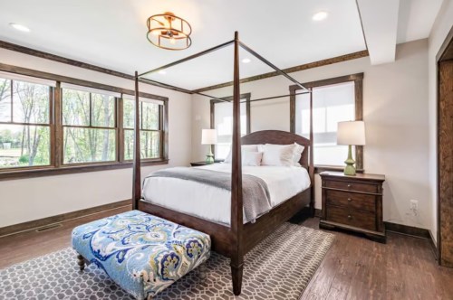 Cozy bedroom featuring a four-poster bed, patterned ottoman, and large windows with natural light.