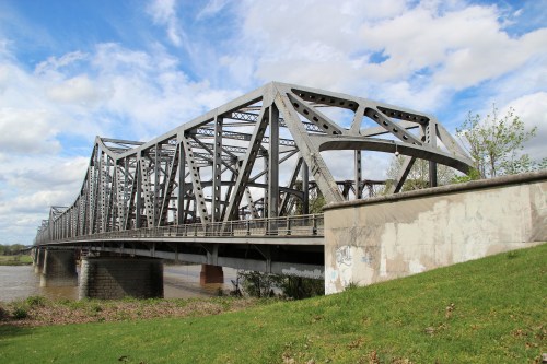 A steel truss bridge spans a river, with green grass in the foreground and a cloudy blue sky above.