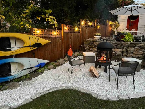 Cozy outdoor space with kayaks, a fire pit, chairs, and decorative lighting surrounded by gravel and greenery.