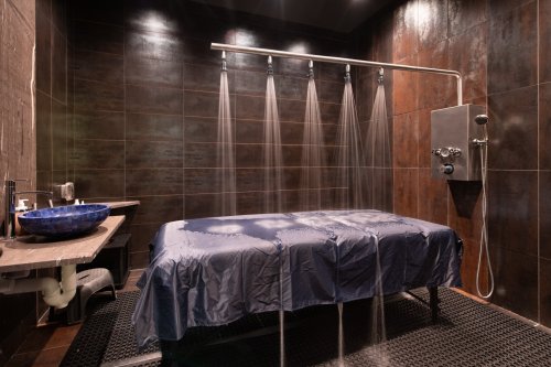 A modern spa room featuring a massage table, overhead shower, and dark tiled walls. Water cascades from multiple showerheads.