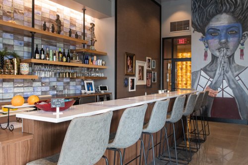 A modern bar with a white countertop, high stools, and a colorful mural of a woman on the wall. Decor includes bottles and art.