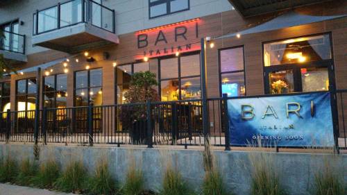 Exterior of Bari Italian restaurant, featuring large windows, outdoor seating, and a sign announcing "Opening Soon."