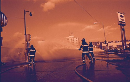 Firefighters spraying water from hoses onto a street, with buildings and a sign in the background.