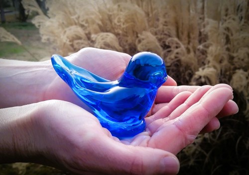A person holds a blue glass bird sculpture in their hands, surrounded by tall grass in the background.