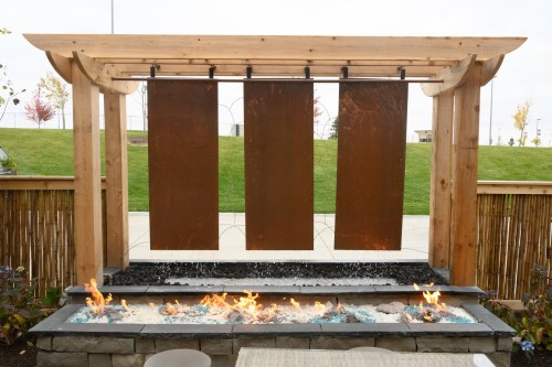 A modern outdoor water feature with three metal panels and a fire pit, surrounded by greenery and stone elements.