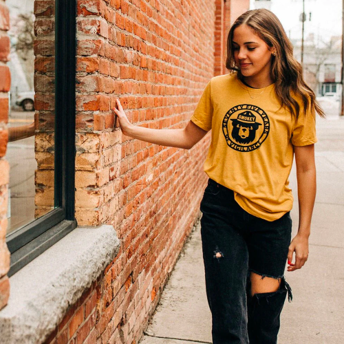 A young woman in a yellow t-shirt and ripped jeans poses against a brick wall, smiling and enjoying the outdoors.