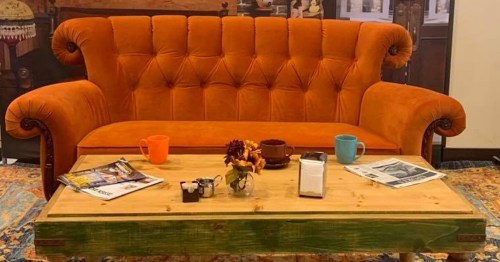 A vibrant orange tufted sofa with a wooden coffee table, featuring mugs, a flower vase, and magazines on a patterned rug.