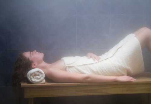 A person relaxes on a wooden bench in a steam room, wrapped in a towel, with a towel under their head.