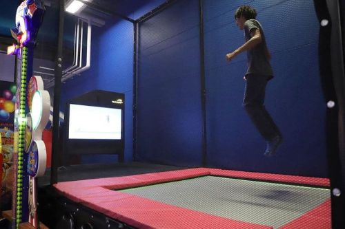 A person jumps on a trampoline inside an indoor play area, with a screen visible in the background.