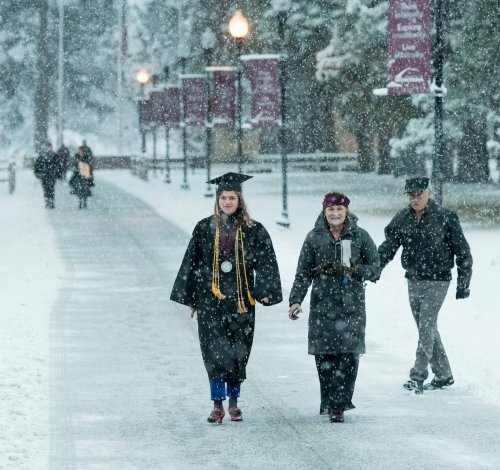 A graduate in a cap and gown walks through falling snow, accompanied by two people on a snowy path.