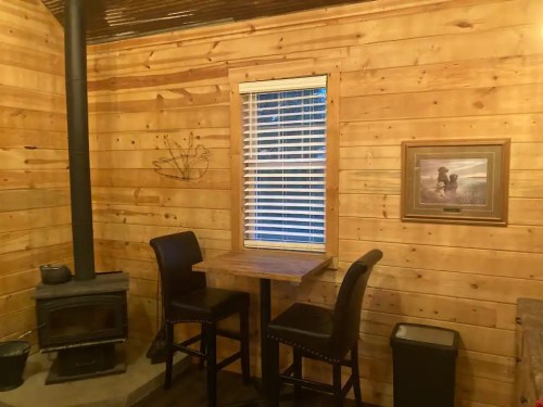 Cozy wooden interior with a small table and chairs, a stove, and a window, featuring rustic decor.