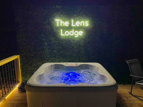 A hot tub illuminated with blue lights, next to a neon sign reading "The Lens Lodge" on a dark wall.