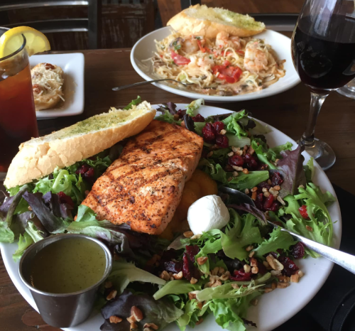 A plate of grilled salmon on a bed of mixed greens with pecans and a side of vinaigrette, accompanied by bread and a drink.
