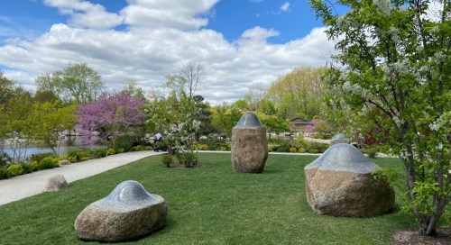 A serene garden scene featuring large, smooth stones, lush greenery, and blooming trees under a cloudy sky.
