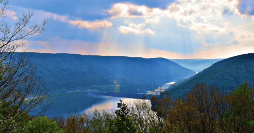 A scenic view of a river winding through lush green hills under a cloudy sky with rays of sunlight breaking through.