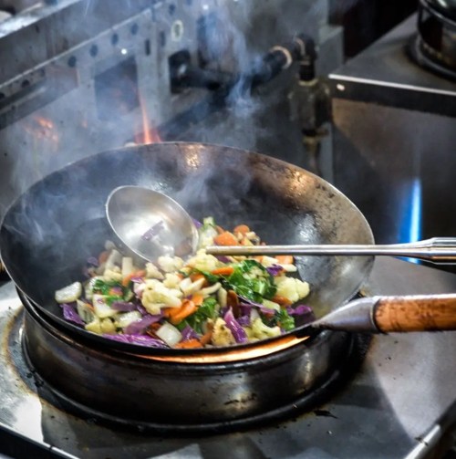 A wok on a stove with colorful vegetables being stir-fried, surrounded by steam and kitchen equipment.