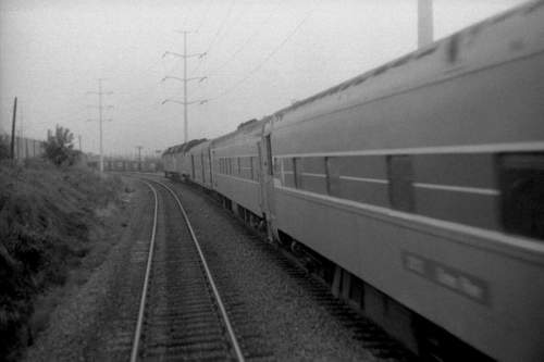 A black and white photo of a train curving along tracks, with power lines in the background and a foggy atmosphere.