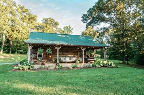 A charming rustic cabin with a green metal roof, surrounded by lush greenery and colorful flowers.