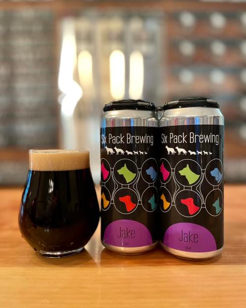 A glass of dark beer next to a four-pack of Six Pack Brewing cans on a wooden table.