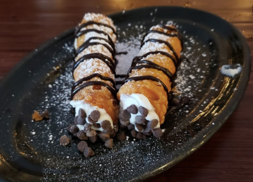 Two cannoli filled with cream, topped with chocolate drizzle and mini chocolate chips, served on a black plate.