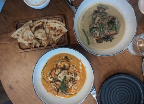 A wooden table with two bowls of curry dishes and a plate of flatbreads. One dish features shrimp, the other beef.
