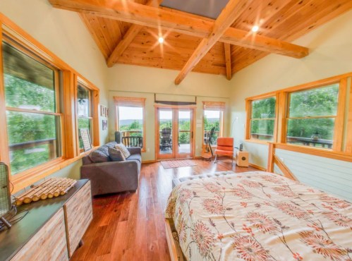 Cozy bedroom with wooden beams, large windows, a bed with floral bedding, and a sitting area with a guitar.