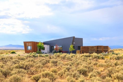 Modern house in a desert landscape, surrounded by sagebrush and distant mountains under a cloudy sky.