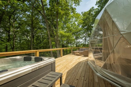 A wooden deck with a hot tub and a geodesic dome, surrounded by lush green trees under a clear blue sky.