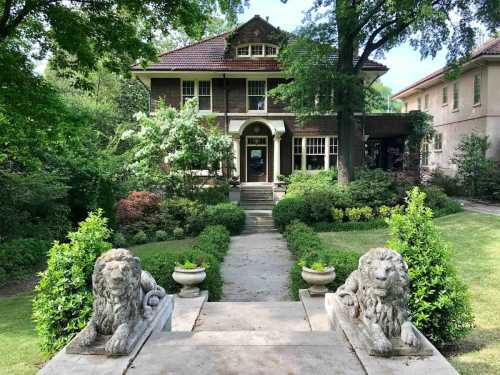 A grand brick house with a porch, surrounded by lush greenery and flanked by stone lion statues at the entrance.