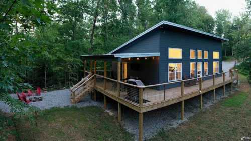 A modern black house with large windows, surrounded by trees, featuring a wooden deck and outdoor seating area.