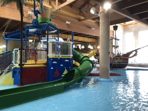Colorful indoor water play area featuring a slide, water features, and a pirate ship in the background.